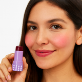 Model holding an open tube of Mo-Beauty Shop's Mo-Jelly Blush + Lip Tint stick in the shade Berry Crush, showing the buildable berry-pink color applied on lips and cheeks, on a white background