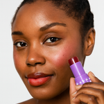 Model holding an open tube of Mo-Beauty Shop's Mo-Jelly Blush + Lip Tint stick in the shade Chili Crisp, showing the buildable red color applied on lips and cheeks, on a white background
