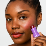 Model holding an open tube of Mo-Beauty Shop's Mo-Jelly Blush + Lip Tint stick in the shade Chili Crisp, showing the buildable red color applied on lips and cheeks, on a white background